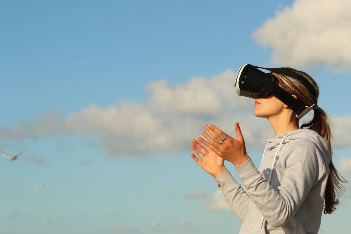 Woman wearing a VR headset outdoors with hands raised against a blue sky