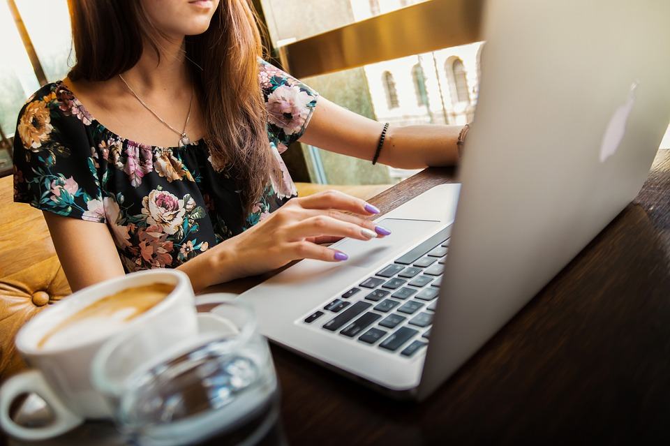 Woman in a floral dress typing on a MacBook laptop at a cafe table with a coffee cup nearby