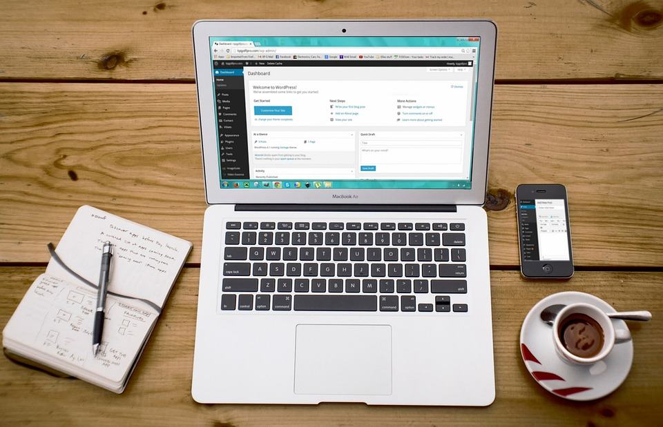 MacBook Air open to a WordPress dashboard on a wooden desk beside a notebook, phone and coffee cup