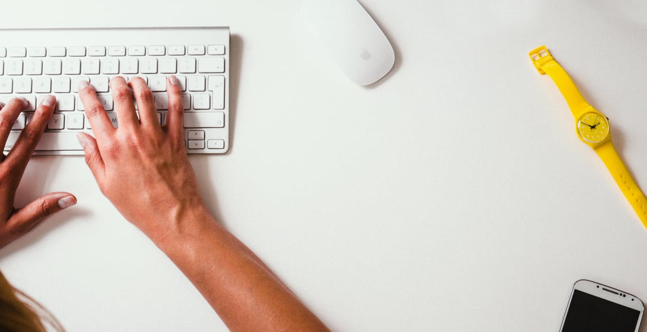 Overhead view of hands typing on a white keyboard with a yellow watch and smartphone on a clean desk