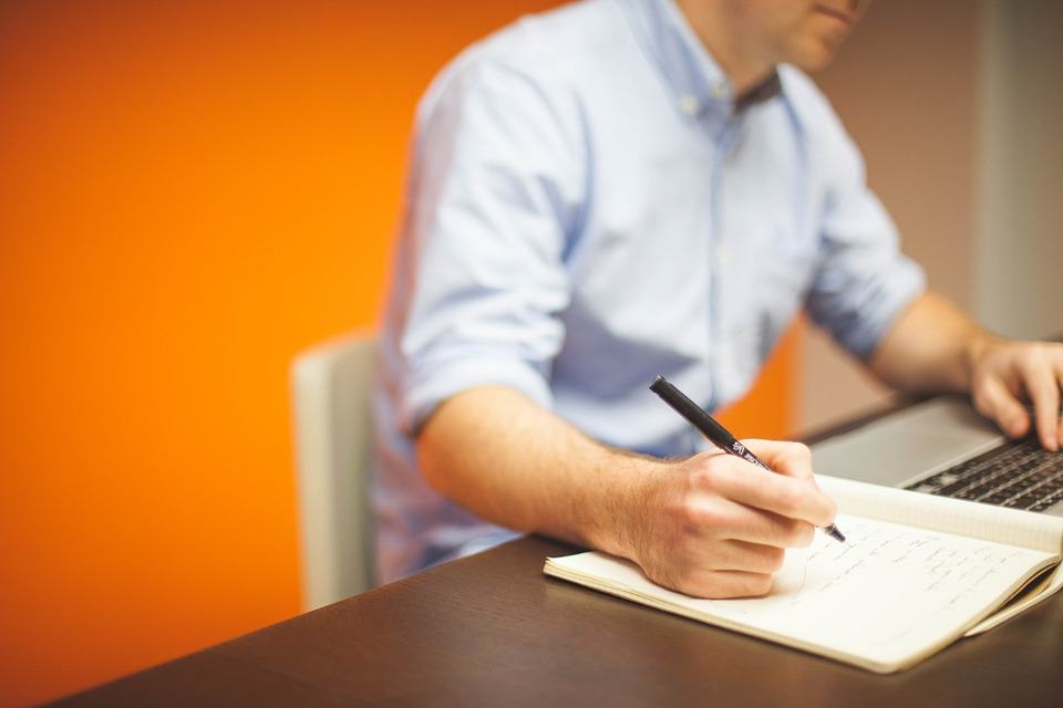 Person in a blue shirt writing notes in a notebook while working on a laptop