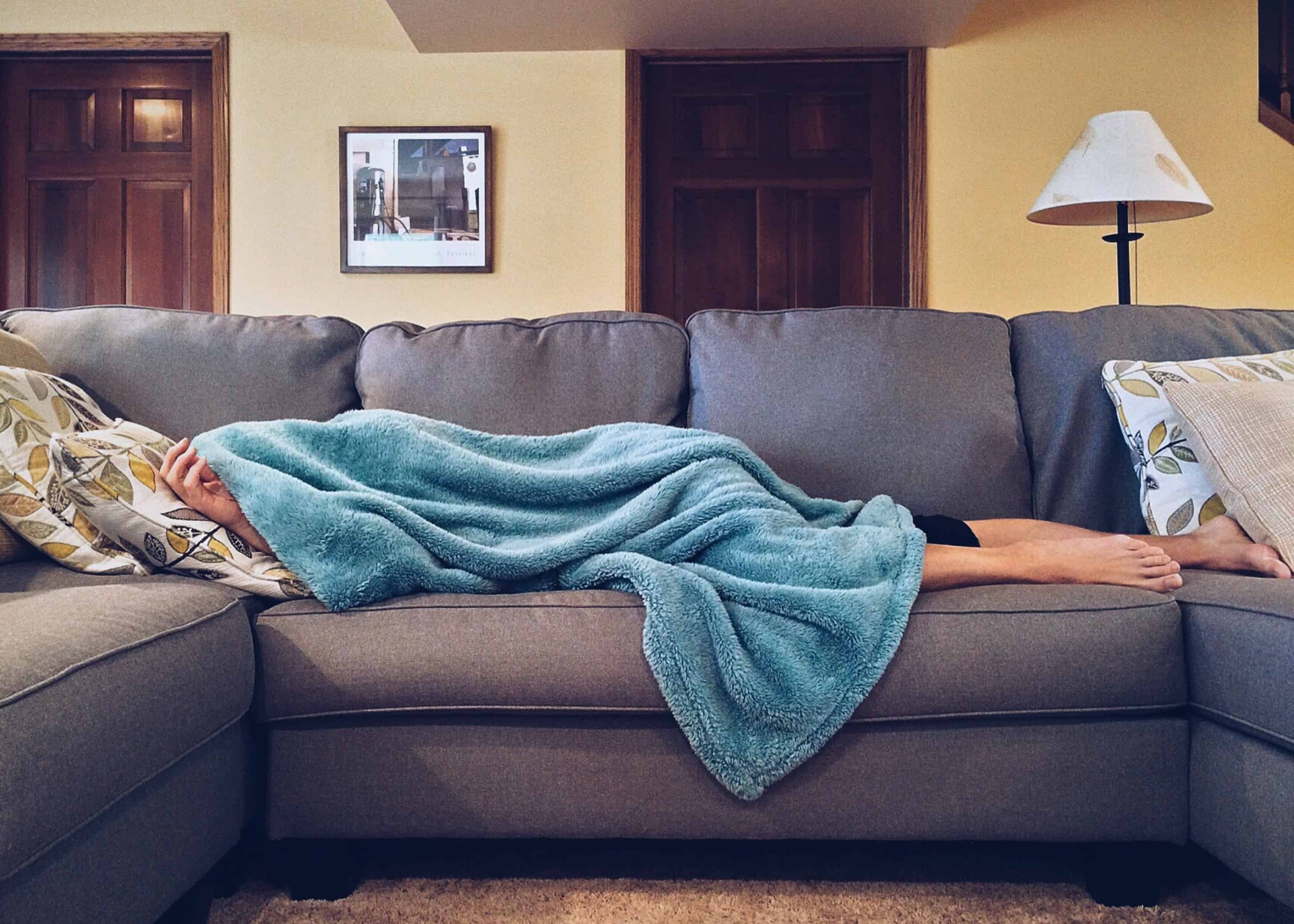 Person lying under a blanket on a grey corner sofa, illustrating procrastination and time management