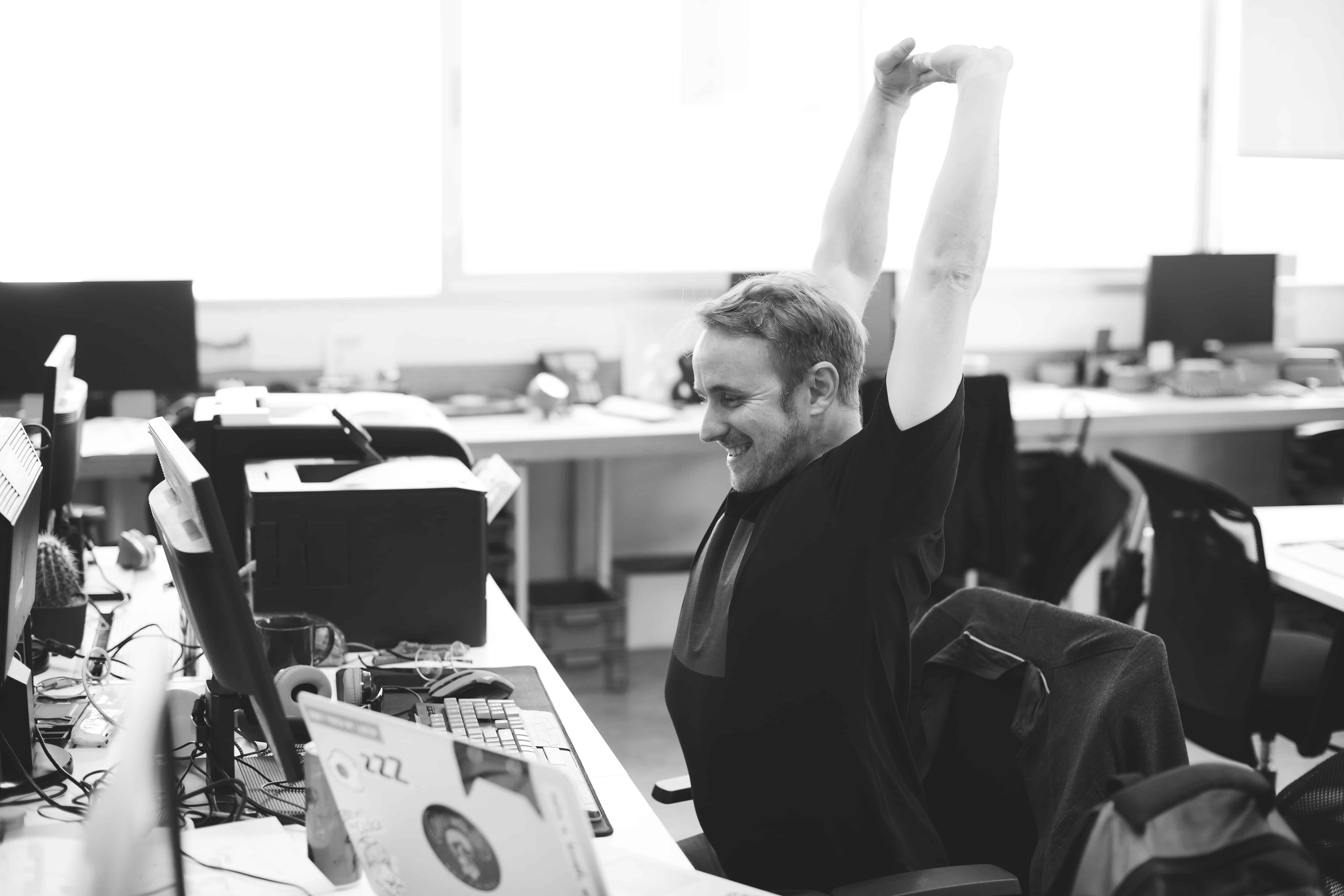Office worker stretching arms above his head and smiling at his desk, illustrating office exercises