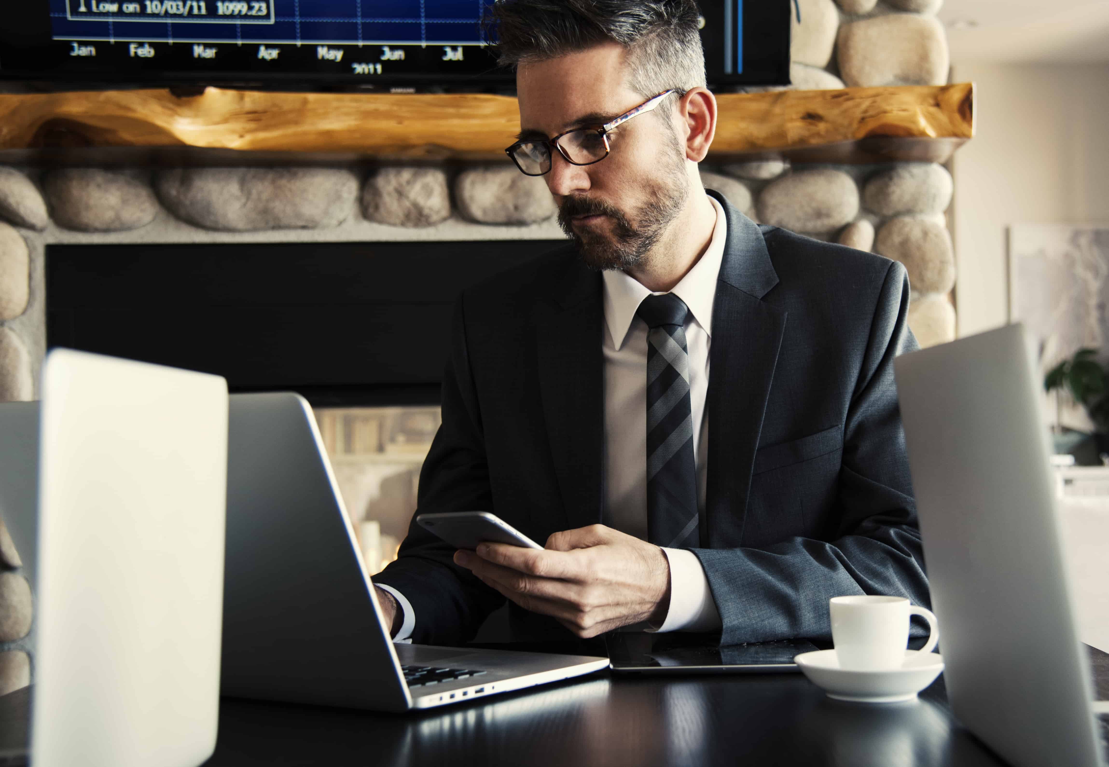 Businessman in a suit checking his smartphone at a desk with two laptops and a coffee, representing digital outsourcing risks