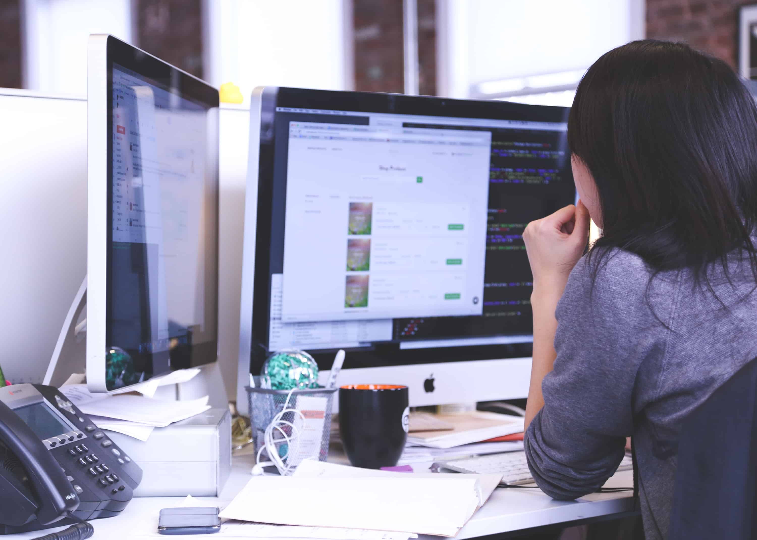 Developer working at two monitors, one showing code and the other a web page, in a busy office environment