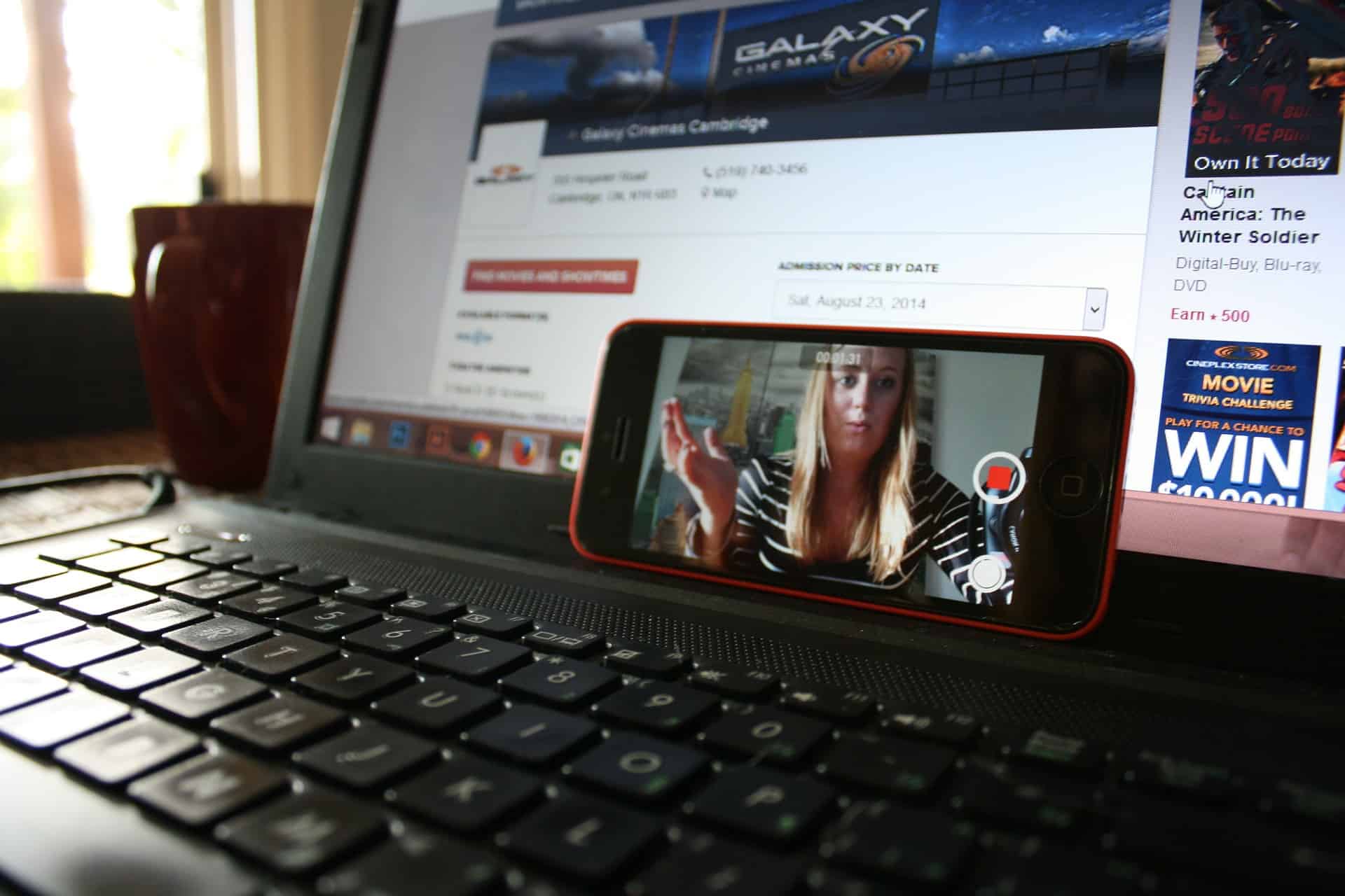 Smartphone propped on a laptop keyboard showing a video recording, with a cinema website visible on the laptop screen