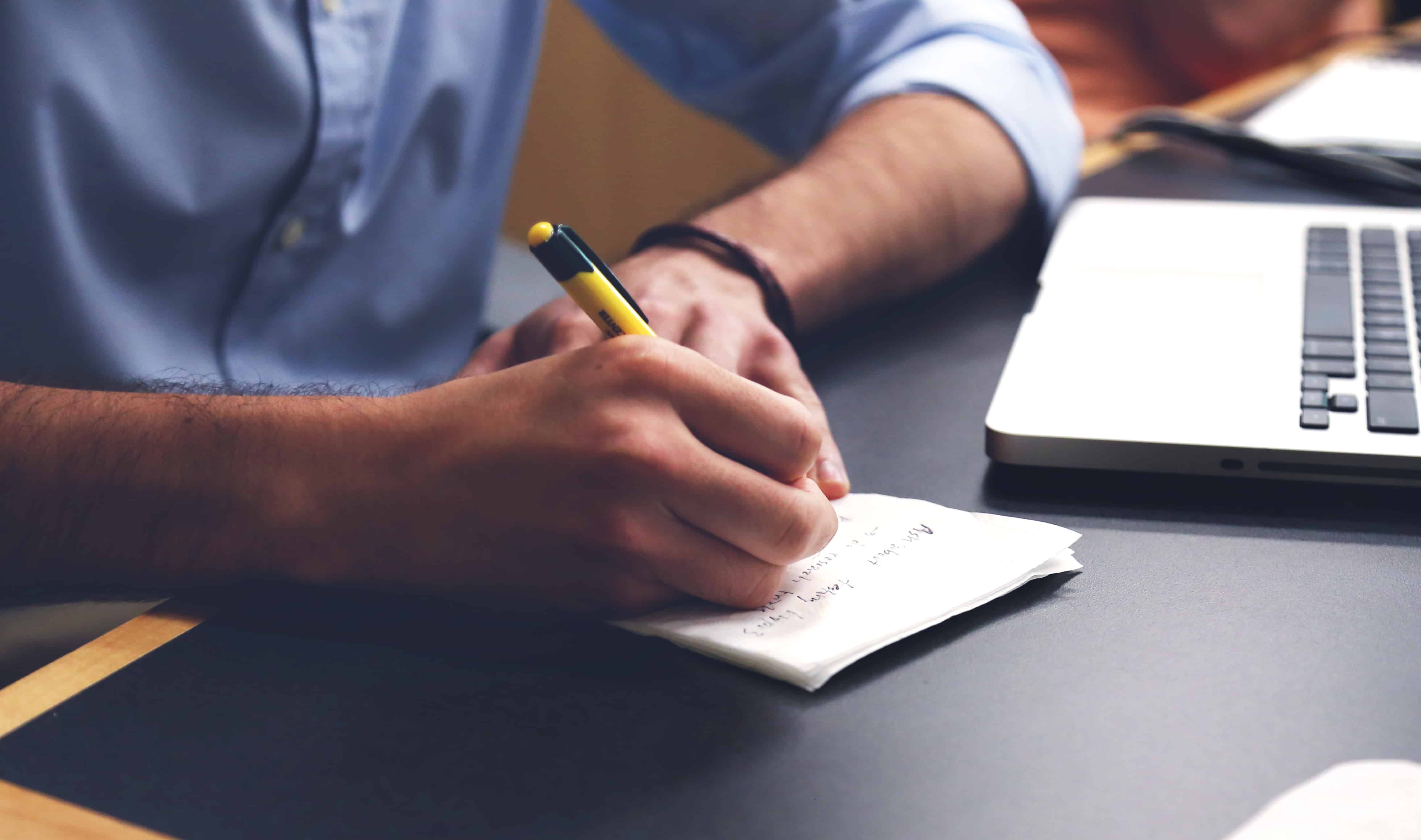 Man writing notes on paper next to a laptop, illustrating an article about duplicate content strategy