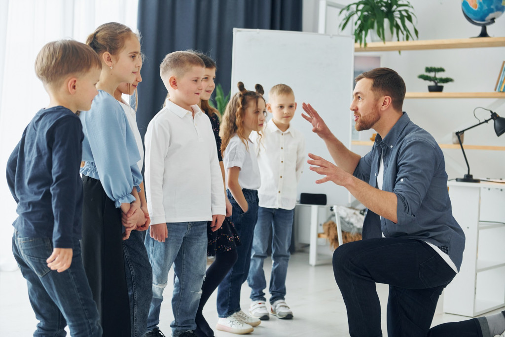Teacher crouching down to speak with a group of young children in a bright classroom